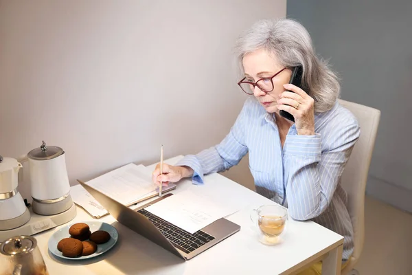 Серьезные Пожилая Женщина Сидит Кухонным Столом Работает Ноутбуком Телефоном Печенье — стоковое фото