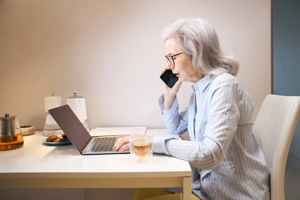 Современная Пожилая Женщина Работает Дома Общается Телефону Кухонном Столе Ноутбука — стоковое фото