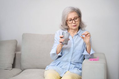 Güzel emekli, elinde bir bardak suyla kanepeye oturmuş hap içiyordu. Yakınlarda bir ilaç kutusu yatıyordu.