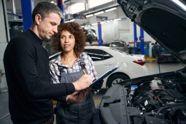 Günlük kıyafetli bir adam araba atölyesinde evrakları imzalıyor. Mavi klasörlü çok kültürlü bir kadın yakınlarda duruyor.