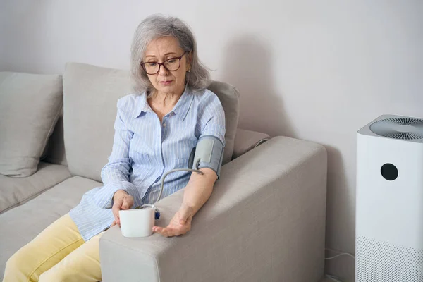 Calm elderly woman sits on the couch and measures blood pressure with a tonometer, she has gray hair