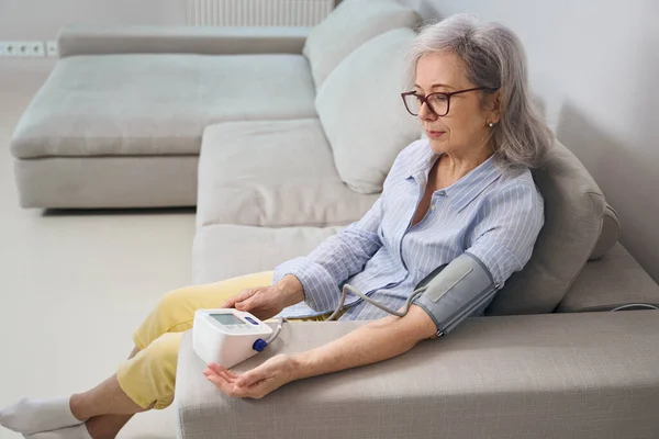소파에 앉아서 혈압을 계량기로 측정하는 나이든 여성은 흰머리입니다 — 스톡 사진