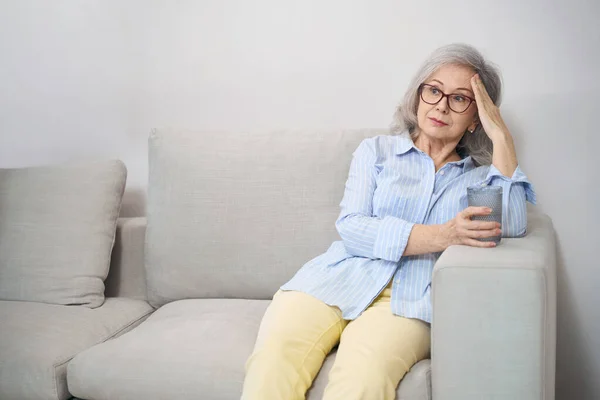 漂亮的退休女人坐在沙发上 喝了一杯水 有点头疼 — 图库照片