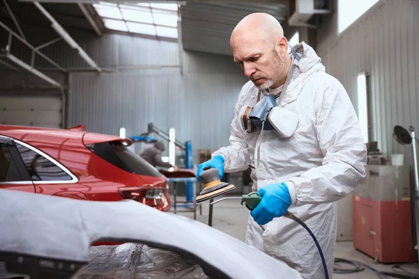 Auto repairman uses a pneumatic tool in the process of grinding and painting a car, a person uses a respirator