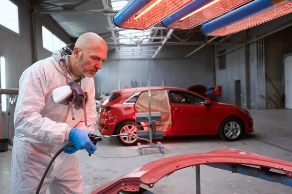 Experienced craftsman in overalls selects paint for a car, he uses a spray gun and paint selection lantern