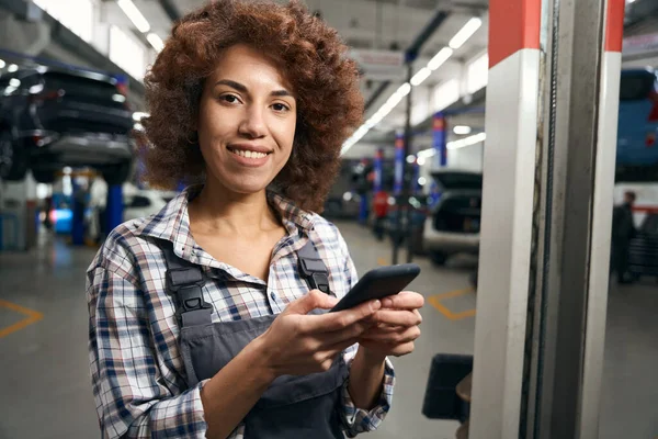 車の修理工場で女性の自動車整備士彼女の手の中に携帯電話を保持します 有料シャツの修理女性 — ストック写真