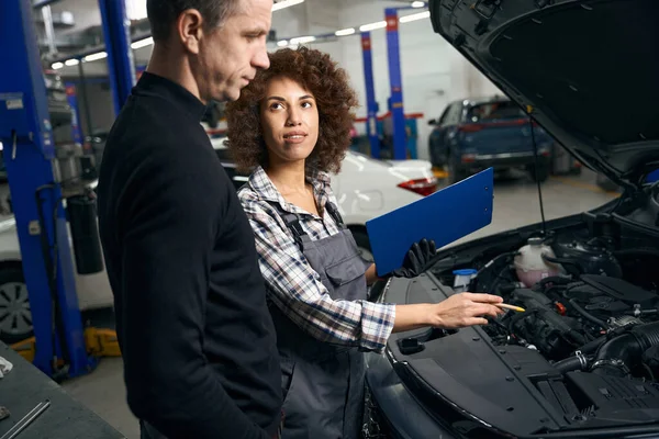 Man in casual clothes communicates with a foreman in an auto repair shop, a multiracial woman holds a blue folder