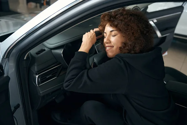 若い女性は新しい車のハンドルに頬を押し当て インテリアは暗い色で作られています — ストック写真