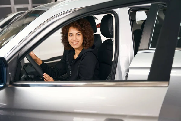 笑顔多人種の女性の買い物客は ブランドの新しい車のサロンでホイールの後ろに座って 彼女は右のモデルを選択します — ストック写真