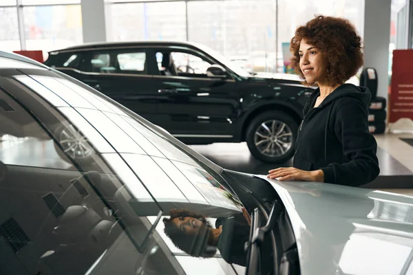 笑顔多人種の女性は 新しい車のフードに彼女の手を置きます 車は専門サロンで販売されています — ストック写真