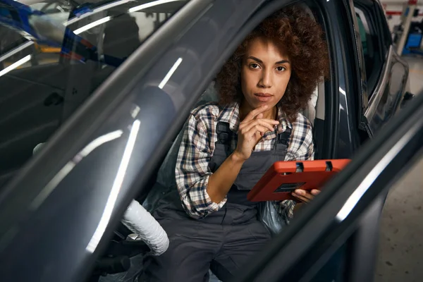 女性の自動車整備士は コンピュータ診断のためのデバイスと車のインテリアに座って ステアリングホイールは汚染から保護されています — ストック写真