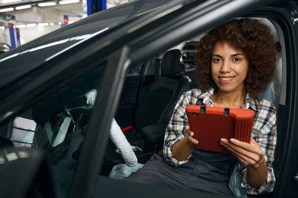 笑顔の女性の自動車整備士は 汚染から保護されたコンピュータ診断機 ステアリングホイールとシートを備えた車の中に座っています — ストック写真