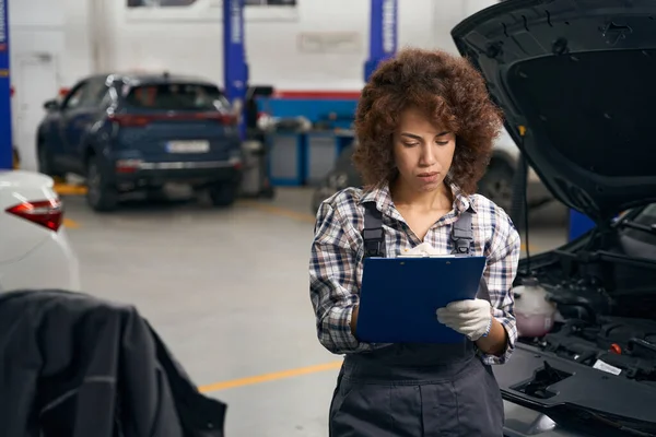 Cute female auto mechanic stands near the car in the workshop, she writes something in a blue folder