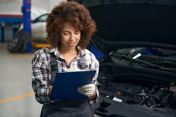 Curly-haired female auto mechanic stands in the middle of a car repair shop, she writes something in a blue folder