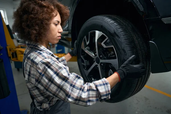 現代の自動車修理工場で予定されているメンテナンス中の車で 女性の修理工は慎重に車のホイールを調べます — ストック写真