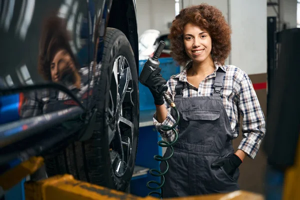 かわいい女性の修理工は 車の車輪で動作するように彼女の手の中に空気圧ドライバを保持し 女性は喜んで笑顔 — ストック写真