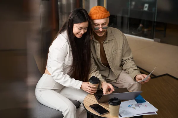 ノートパソコンの画面で笑顔の女性同僚に何かを示す机に座って手に文書を持つ企業の従業員 — ストック写真