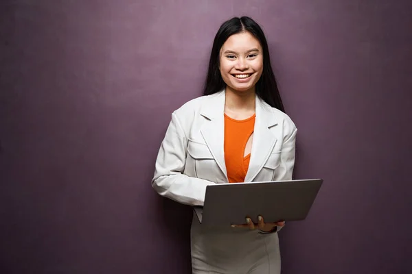 Smiling happy elegant lady with laptop in hands standing against wall and looking before her