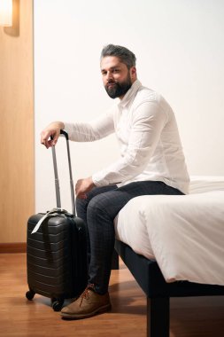 Bavulları otel odasında yatağın kenarında oturan sakin bir adam.