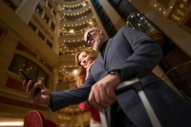 Yetişkin bir adam ve güzel bir kadın otelin lobisinde, lüks bir iç mekanda selfie çekiyorlar.