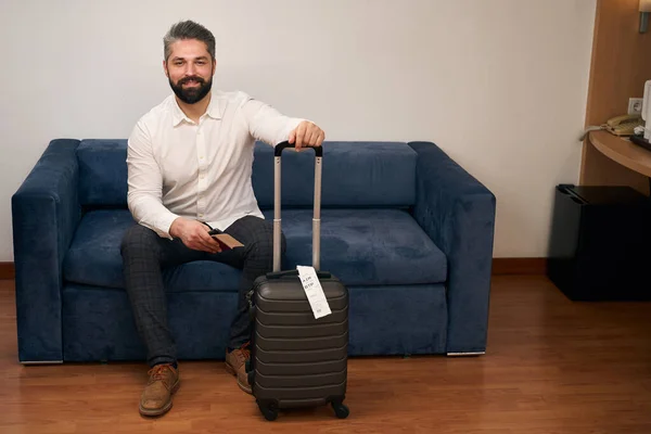 Zufriedener Mann Mit Rollkoffer Und Reisepass Sitzt Auf Sofa Hotelzimmer — Stockfoto