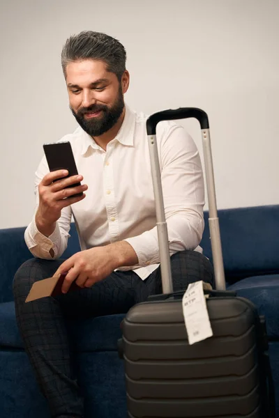 持旅行证件的心满意足的男性创业者坐在酒店房间的沙发上 看着智能手机屏幕 — 图库照片