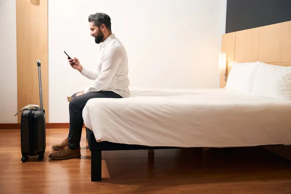 坐在床边的带着智能手机的愉快游客的侧视图 — 图库照片