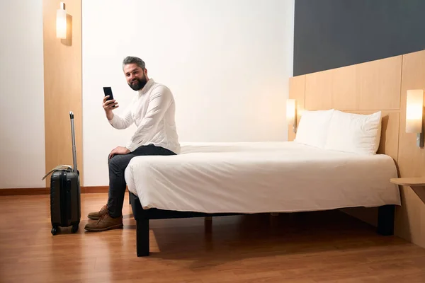喜びの男とともに携帯電話で手に座っているエッジのベッドでホテルの部屋 — ストック写真