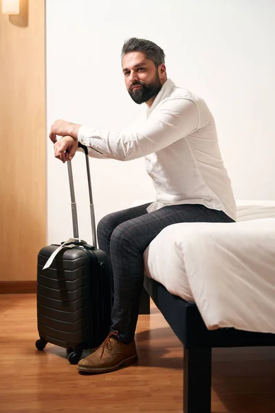 トロリースーツケースを着た思慮深い紳士がホテルの部屋のベッドの端に座って — ストック写真