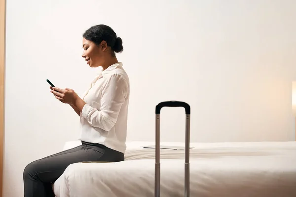 手持手机坐在豪华套间床上微笑的女企业家的侧影 — 图库照片