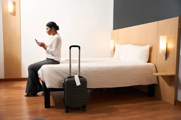 ホテルの部屋でベッドの端に座って手に携帯電話で集中女性観光客の側面図 — ストック写真