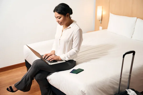 Concentrated stylish lady seated on bed in hotel room typing on laptop