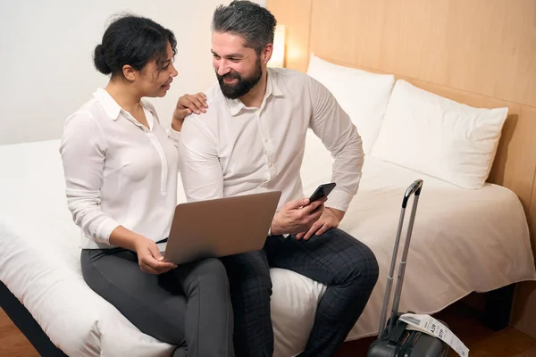 stock image Cheerful man with smartphone and smiling woman with laptop sitting on bed in hotel room