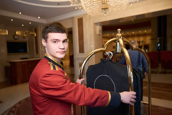 Young bellboy is carrying a trolley with luggage through the lobby, in a luxury design hotel