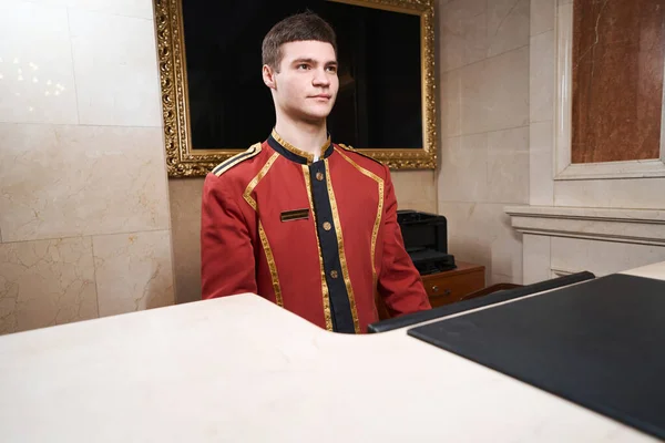 Young Pleasant Bellhop Standing Reception Desk Guy Traditional Uniform — Stock Photo, Image
