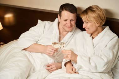 Orta yaşlı erkek ve kadın şampanyayı yatakta içerken çift büyük bir yatağa yerleşti.