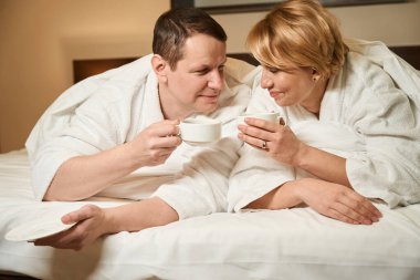 Orta yaşlı kadın ve bornozlu kocası yatakta kahve içiyorlar, çift rahat bir otel odasında kalıyor.