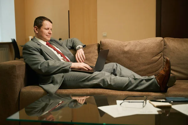 ビジネススーツの男はノートパソコンで働きビジネスマンは足のある居心地の良いソファに位置しています — ストック写真