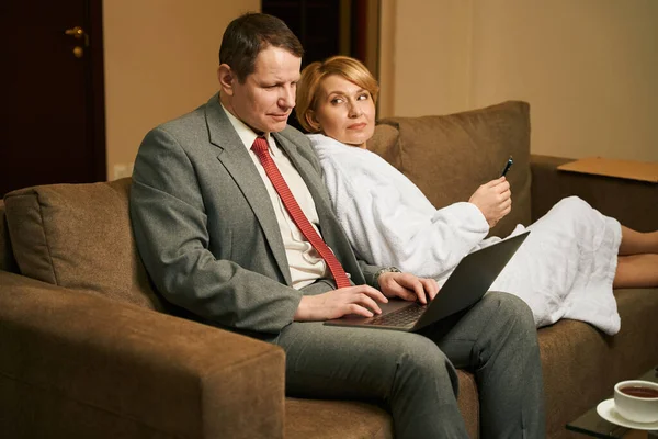 ビジネススーツの男性は柔らかいソファのラップトップで動作し 彼の妻は携帯電話で彼の隣に座っています — ストック写真