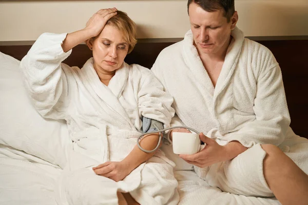 男はベッドの上に座り 気圧計で妻の圧力を測定し 女性は頭痛を持っています — ストック写真