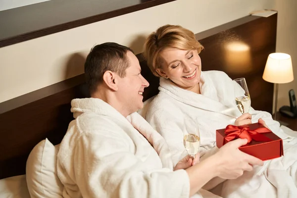 幸せな夫は赤い箱で彼の最愛の妻のギフトを与えます カップルはベッドの中でシャンパンで彼らの結婚記念日を祝います — ストック写真