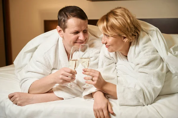 かわいい中年の配偶者はベッドでシャンパンを飲む カップルは快適なベッドルームに位置しています — ストック写真