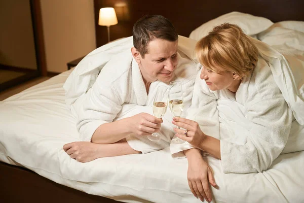中年の女性とバスローブの彼女の夫はベッドでシャンパンを飲んでいる カップルは快適なベッドルームに位置しています — ストック写真