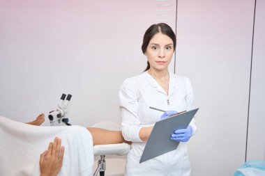 Kadın jinekolog ayakta duruyor ve klinikteki hastanın şikayetlerini yazıyor.