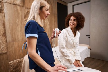 Güzel bir kadın spa salonundaki bir kanepede oturuyor ve bir RF kaldırma uzmanıyla konuşuyor.