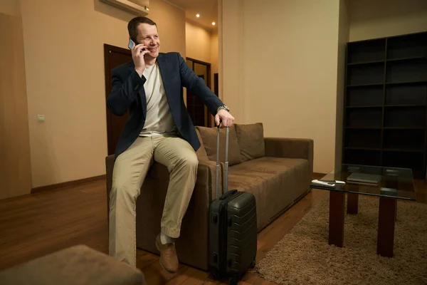 旅行用のスーツケースを着た笑顔の男がホテルの部屋に座って 彼は携帯電話で通信します — ストック写真