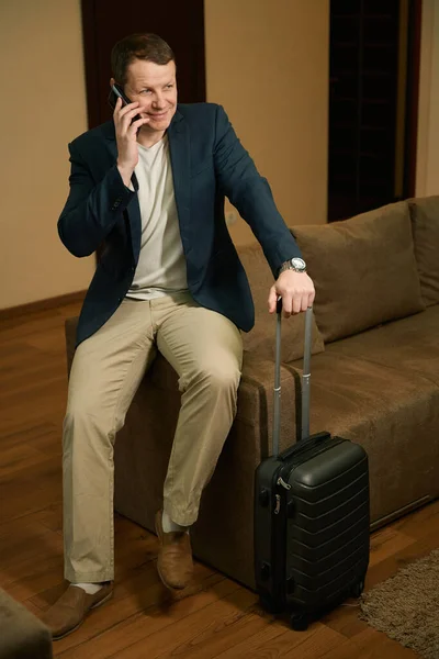 一个带着旅行箱愉快地旅行的男人正坐在宾馆房间里 他在用手机交谈 — 图库照片