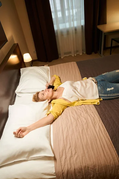旅行服の美しい女性は大きなベッドの上に横たわっている 彼女の腕を伸ばし 彼女は電話で話している — ストック写真