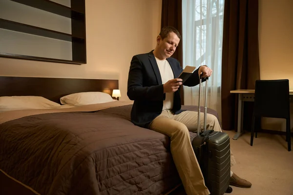 穿着旅行服装的男人坐在宾馆房间的一张大床上 手里拿着护照和车票 — 图库照片