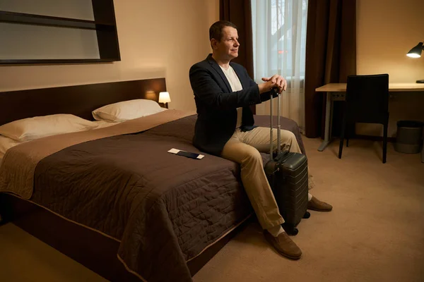 穿着旅行服装的男人坐在旅馆房间的卧室里 前面是一个旅行行李箱 — 图库照片